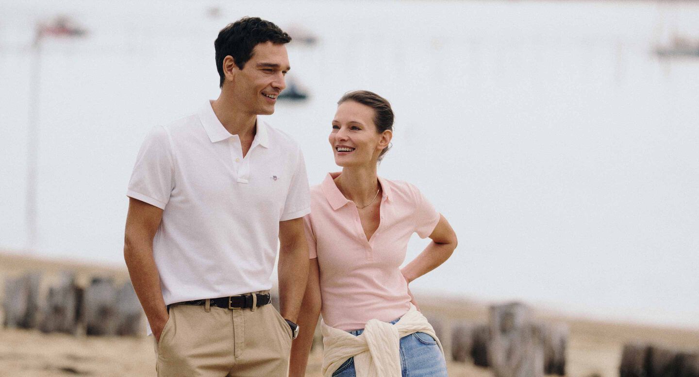 Gant polo shirts worn by two people walking along a beach, one in white and one in light pink.
